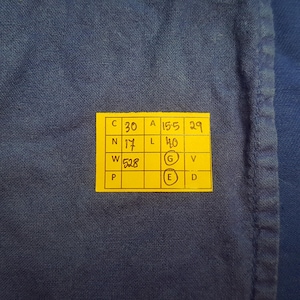 May include: Close-up of a textured, dark blue fabric with a bright yellow tag. The tag has black text and numbers, including "30", "15.5", "29", "17", and "40". The fabric is a solid color, and the tag is rectangular.
