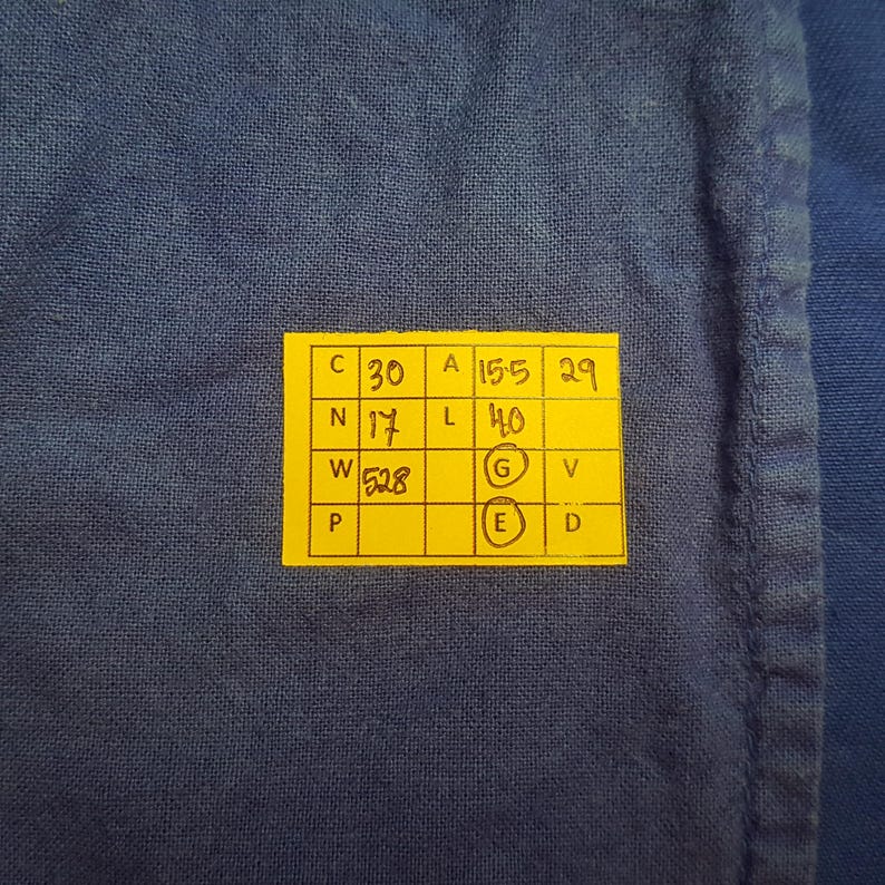 May include: Close-up of a textured, dark blue fabric with a bright yellow tag. The tag has black text and numbers, including "30", "15.5", "29", "17", and "40". The fabric is a solid color, and the tag is rectangular.