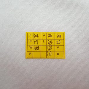 May include: A close-up of a yellow rectangular tag with handwritten black numbers and letters. The tag is on a white fabric background. The numbers include 27, 24, 24, 19, 26, 23, 418.