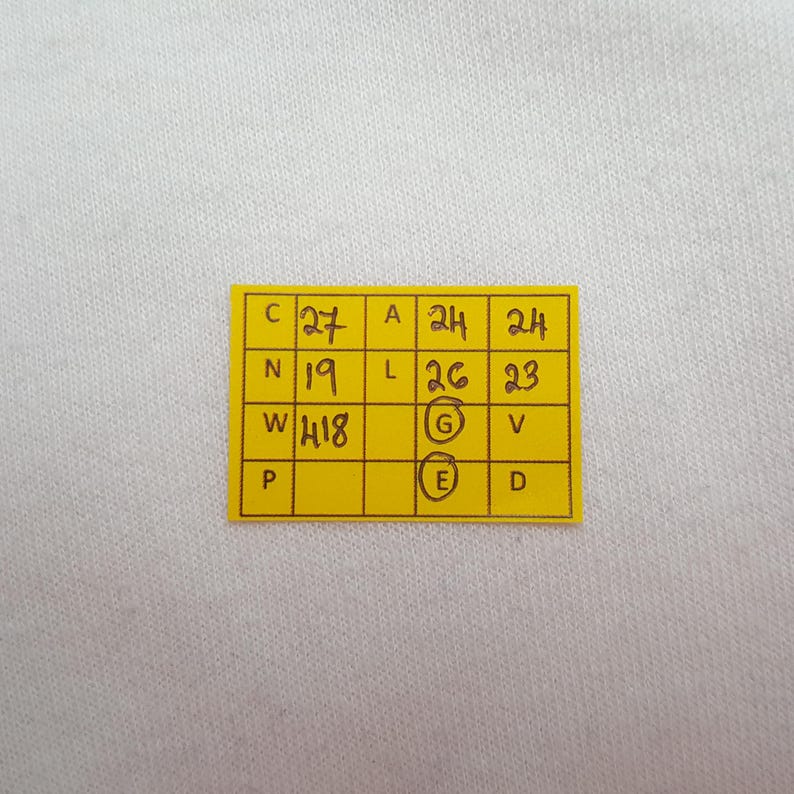 May include: A close-up of a yellow rectangular tag with handwritten black numbers and letters. The tag is on a white fabric background. The numbers include 27, 24, 24, 19, 26, 23, 418.
