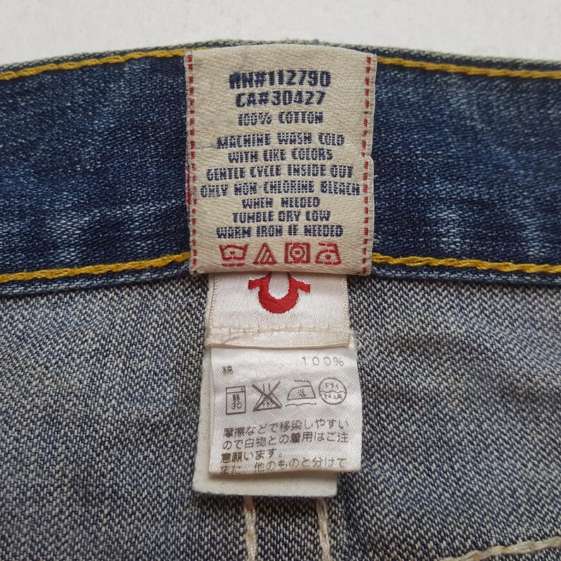 May include: Close-up of a denim garment tag with text including "AN#112790", "CA#30427", and washing instructions. The tag is white and red, with a horseshoe logo. The garment is made of 100% cotton.
