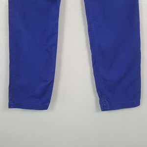 May include: Close-up of the lower portion of a pair of royal blue pants. The pants are made of a solid, smooth fabric and have visible stitching along the seams. The image is set against a plain, light-colored background.