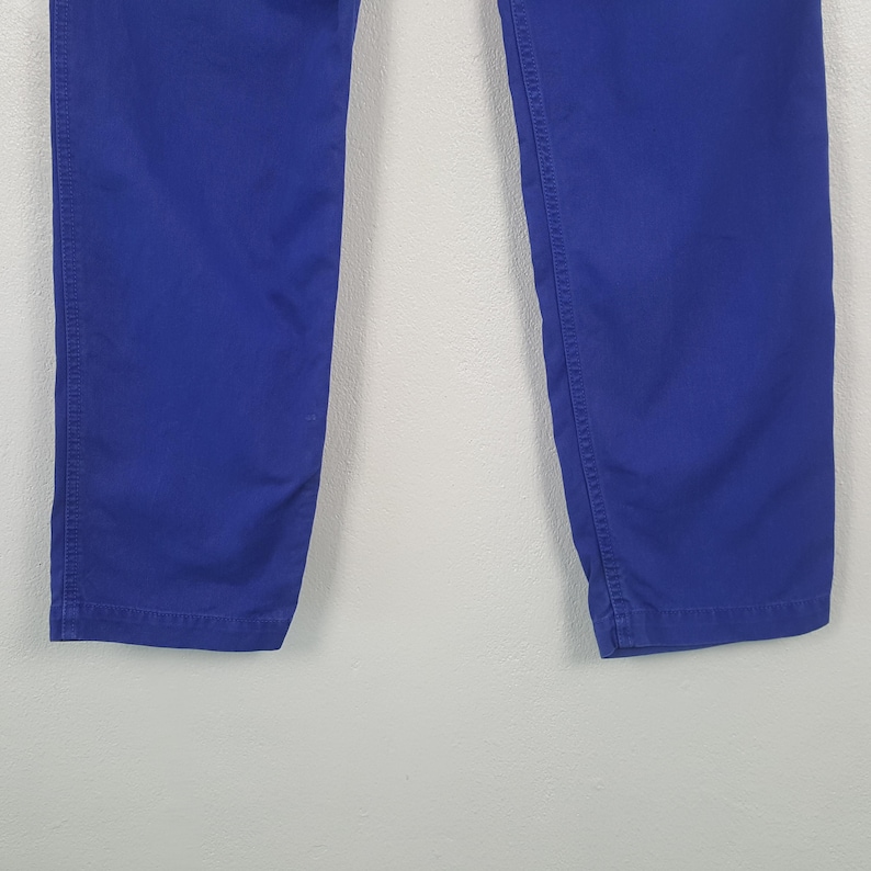 May include: Close-up of the lower portion of a pair of royal blue pants. The pants are made of a solid, smooth fabric and have visible stitching along the seams. The image is set against a plain, light-colored background.