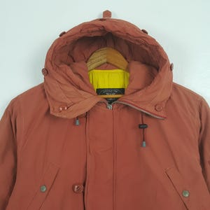 May include: A rust-colored hooded jacket with a zipper closure and multiple button details. The jacket features a high collar and a yellow lining visible inside the hood. The jacket is hanging on a wooden hanger.