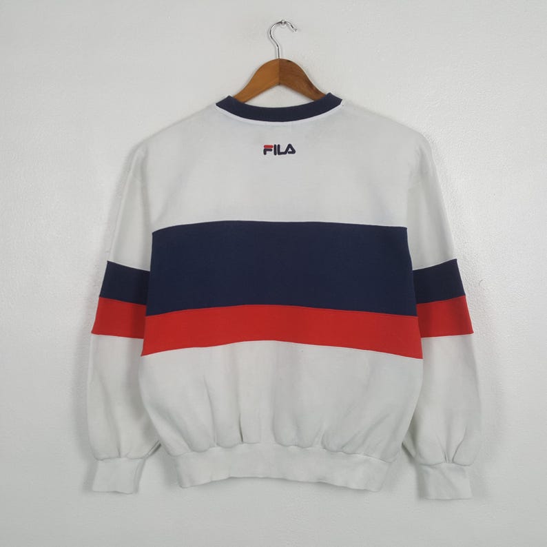 May include: White sweatshirt with navy blue and red horizontal stripes across the chest. The long-sleeved top has a crew neck and the brand name "FILA" is embroidered in red on the upper back. The sweatshirt is hanging on a wooden hanger.