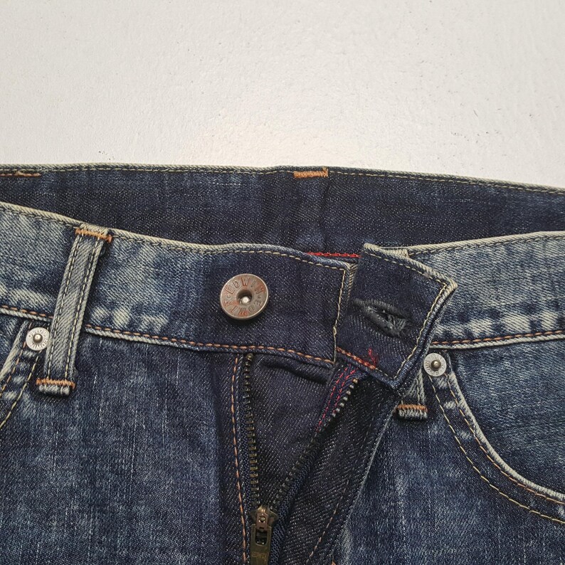 May include: Close-up of a pair of blue denim jeans. The image shows the button, zipper, and stitching details. The jeans have a classic five-pocket design with a metal button closure and a zipper fly.