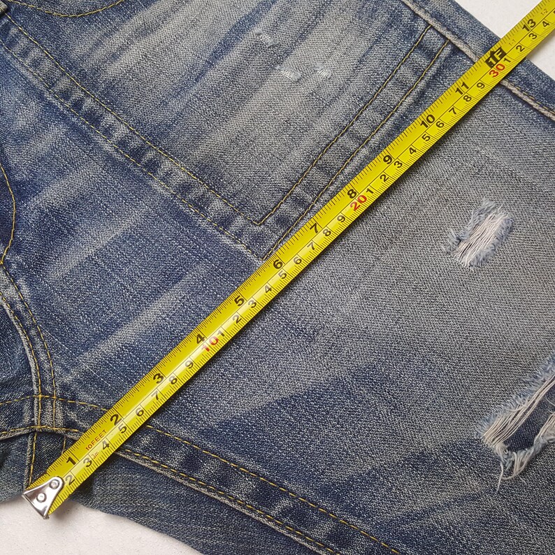 May include: Close-up of blue denim jeans with distressed details. A yellow tape measure shows measurements in inches. The jeans have visible stitching and a worn appearance, with a rip near the leg.