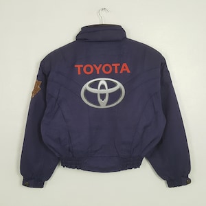 May include: Navy blue Toyota jacket with a silver logo and red "TOYOTA" text on the back. The jacket has a high collar, a brown patch on the left shoulder, and a ribbed hem. The jacket is hanging on a metal hanger.