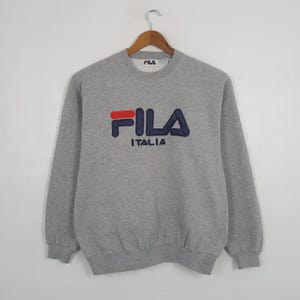 May include: A gray FILA Italia sweatshirt with a crew neck and long sleeves. The FILA logo is embroidered in red and navy blue. The sweatshirt is hanging on a wooden hanger against a white background.