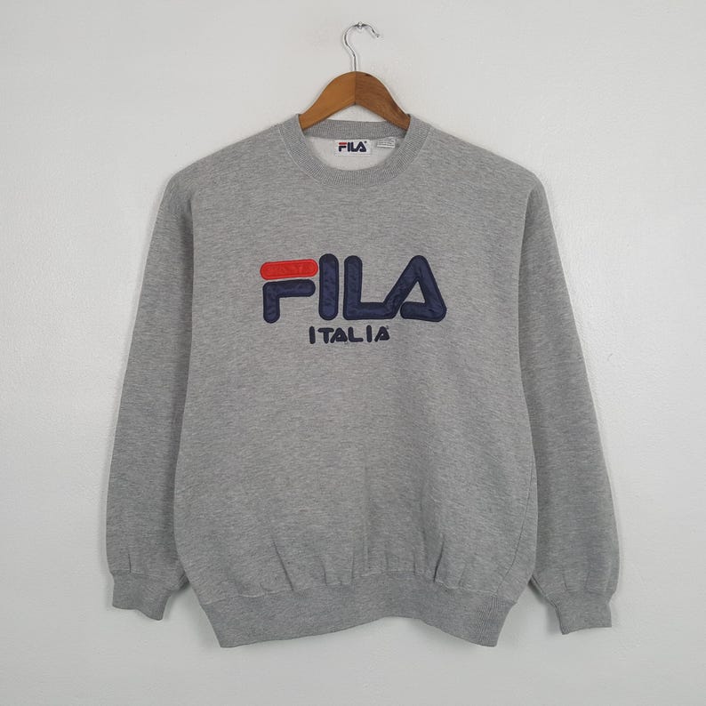 May include: A gray FILA Italia sweatshirt with a crew neck and long sleeves. The FILA logo is embroidered in red and navy blue. The sweatshirt is hanging on a wooden hanger against a white background.