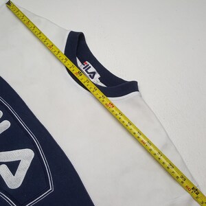 May include: White Fila sweatshirt with navy blue accents. The sweatshirt features a navy blue collar and a large navy blue panel on the left side with the Fila logo. A measuring tape is laid across the sweatshirt.