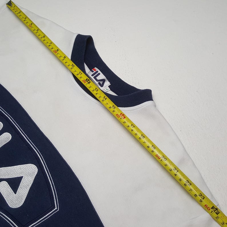 May include: White Fila sweatshirt with navy blue accents. The sweatshirt features a navy blue collar and a large navy blue panel on the left side with the Fila logo. A measuring tape is laid across the sweatshirt.