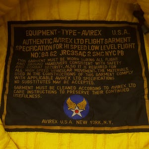 May include: A close-up of a black label with gold trim from an Avirex flight garment. The label includes text such as "AUTHENTIC AVIREX LTD FLIGHT GARMENT" and "AVIREX U.S.A. NEW YORK, N.Y." A circular patch with a star and wings is also visible.