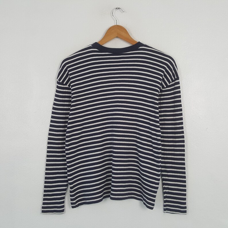 May include: A long-sleeved shirt with horizontal navy blue and white stripes. The shirt is hanging on a wooden hanger against a white background. The shirt has a crew neck and a relaxed fit.