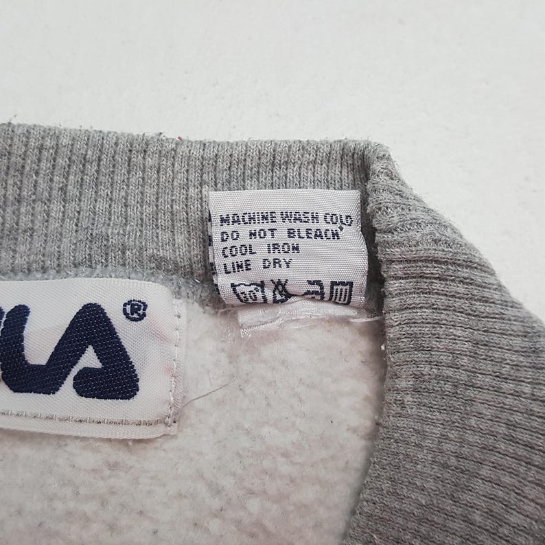 May include: Close-up of a gray sweatshirt collar with a white tag. The tag has washing instructions and a Fila logo. The text on the tag reads: Machine wash cold, do not bleach, cool iron, line dry.