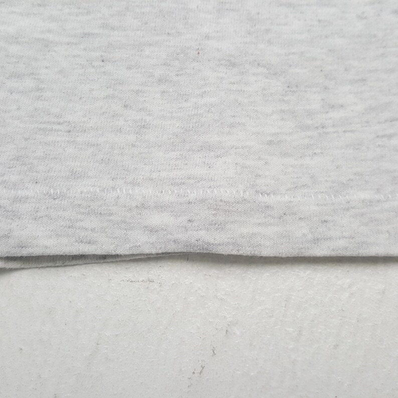 May include: Close-up of a light gray fabric with a subtle speckled pattern. The fabric is folded at the bottom, revealing a seam with visible stitching. The texture appears soft and slightly textured, suggesting a comfortable material.