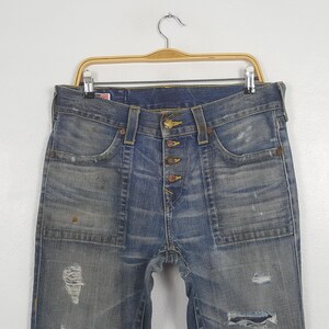 May include: Blue denim jeans with a button fly and distressed details, including rips and faded patches. The jeans have front pockets and are hanging on a wooden hanger.
