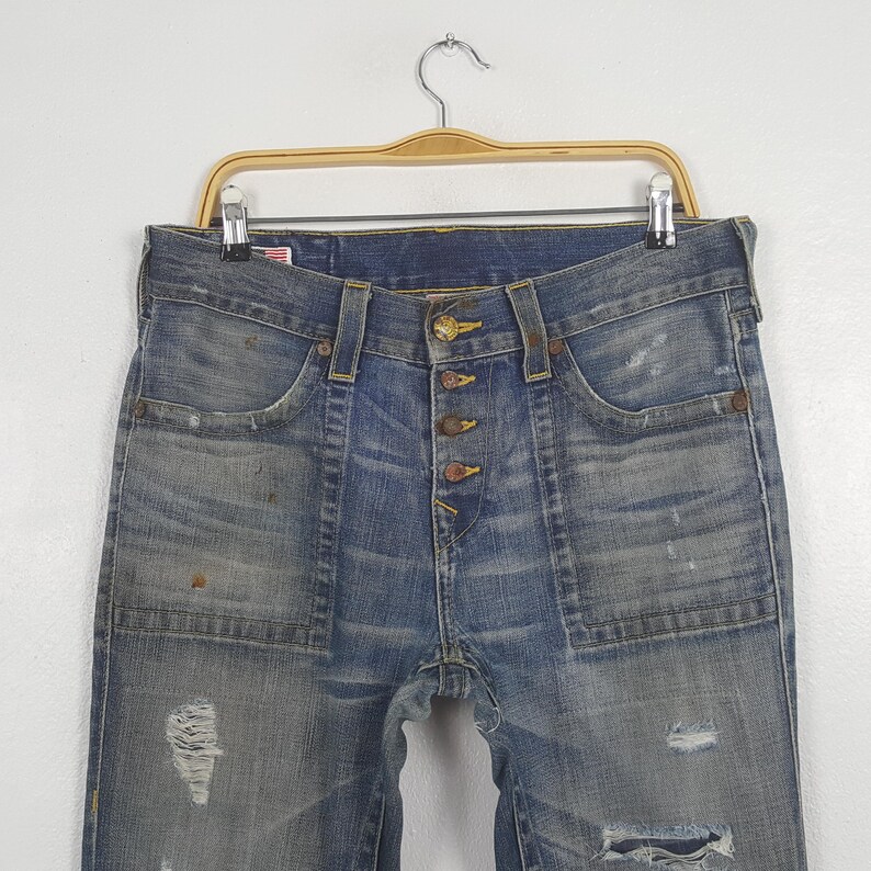 May include: Blue denim jeans with a button fly and distressed details, including rips and faded patches. The jeans have front pockets and are hanging on a wooden hanger.