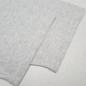 May include: Close-up of a light gray fabric, possibly a t-shirt or other garment. The fabric is folded, revealing a seam. The material appears soft and slightly textured, with a subtle heathered effect. The overall look is simple and minimalist.
