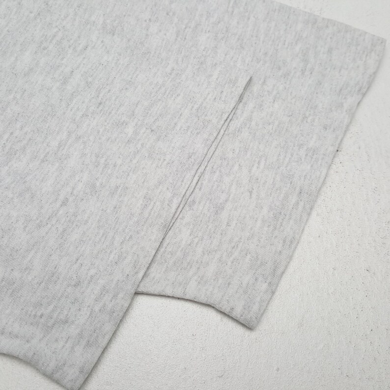 May include: Close-up of a light gray fabric, possibly a t-shirt or other garment. The fabric is folded, revealing a seam. The material appears soft and slightly textured, with a subtle heathered effect. The overall look is simple and minimalist.