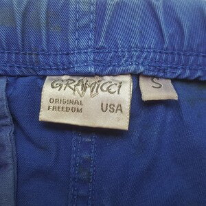 May include: Close-up of blue fabric with a Gramicci label. The label reads "GRAMICCI", "ORIGINAL FREEDOM", and "USA". A small "S" tag is also visible. The fabric has a textured appearance.