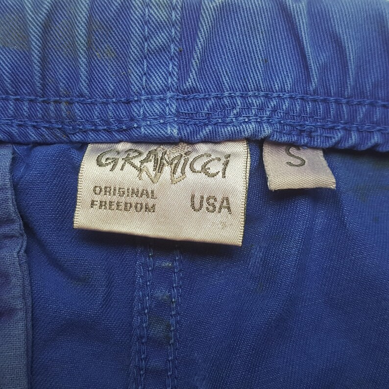 May include: Close-up of blue fabric with a Gramicci label. The label reads "GRAMICCI", "ORIGINAL FREEDOM", and "USA". A small "S" tag is also visible. The fabric has a textured appearance.