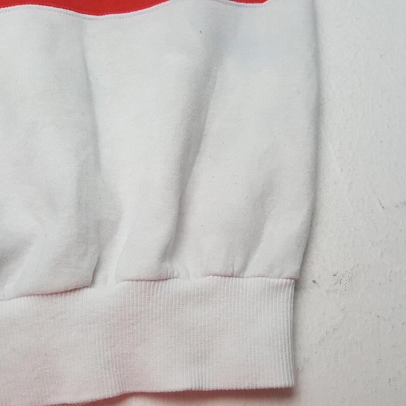May include: Close-up of a white and red sweatshirt. The top portion is red, transitioning to white. The bottom hem and cuffs are ribbed white fabric. The fabric appears soft and slightly textured.