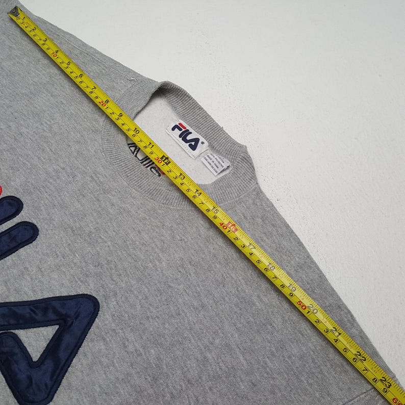 May include: A gray Fila sweatshirt with a crew neck and long sleeves. The Fila logo is prominently displayed in navy blue and red on the front. A yellow tape measure is laid across the sweatshirt.