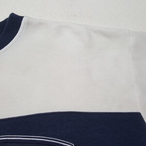 May include: Close-up of a white and navy blue sweatshirt. The top portion is white, with a navy blue band across the lower section. The neckline is navy blue. The fabric appears to be a soft, knit material.