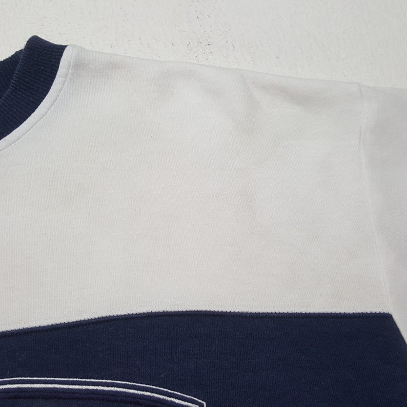May include: Close-up of a white and navy blue sweatshirt. The top portion is white, with a navy blue band across the lower section. The neckline is navy blue. The fabric appears to be a soft, knit material.
