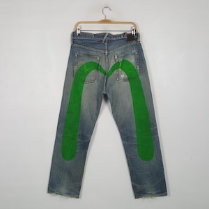 Puede incluir: Vaqueros de mezclilla azul con un gran gráfico de gaviota pintado en verde brillante sobre los bolsillos traseros y las piernas. Los pantalones tienen un diseño clásico de cinco bolsillos, cierre de botones y un lavado desgastado.