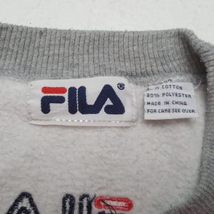May include: Close-up of a gray sweatshirt with a white tag featuring the FILA logo in red and navy blue. The tag also includes text indicating the fabric composition and origin. The sweatshirt has a gray collar.
