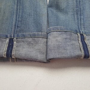 May include: Close-up of the folded cuffs of blue denim jeans. The cuffs are turned up, revealing a darker blue selvedge edge with white stitching. The denim has a faded, worn appearance.