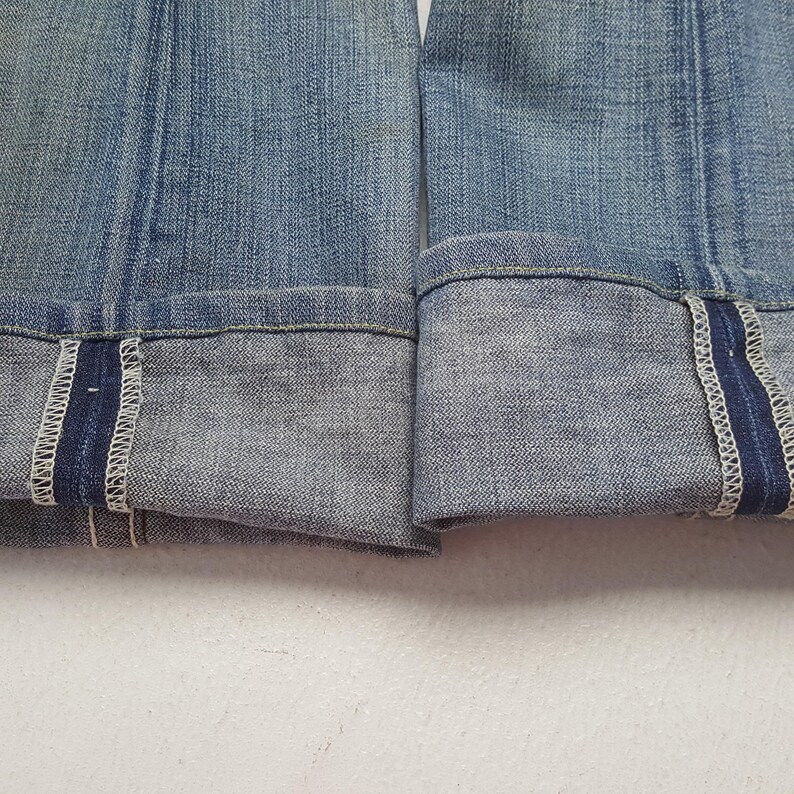 May include: Close-up of the folded cuffs of blue denim jeans. The cuffs are turned up, revealing a darker blue selvedge edge with white stitching. The denim has a faded, worn appearance.