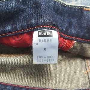 May include: Close-up of a pair of blue denim jeans with a red interior lining. The tag reads "EDWIN" with the numbers "53598" and size "M". Additional codes are printed below the size.