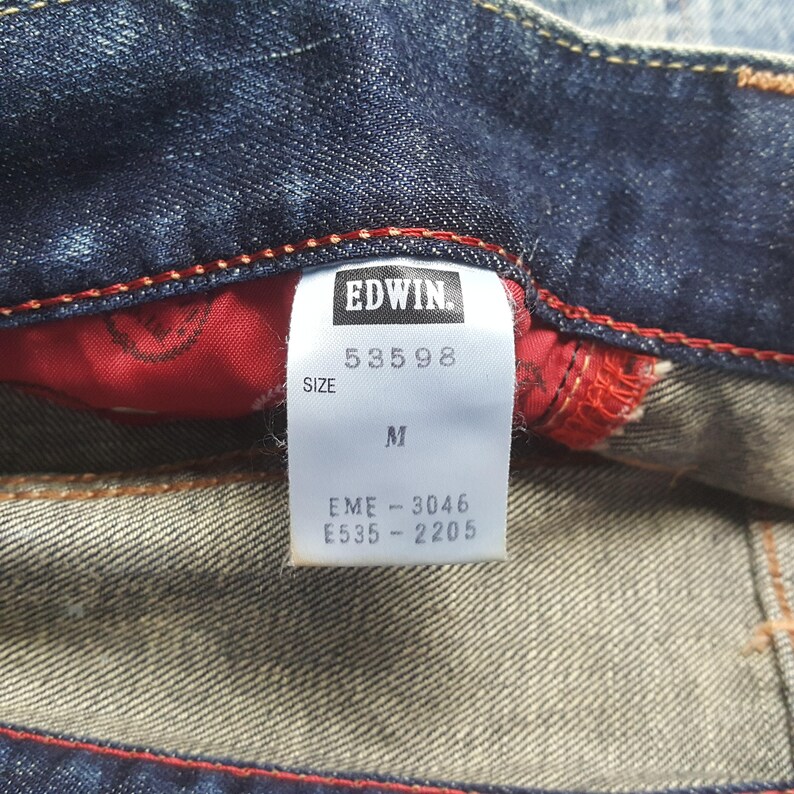 May include: Close-up of a pair of blue denim jeans with a red interior lining. The tag reads "EDWIN" with the numbers "53598" and size "M". Additional codes are printed below the size.