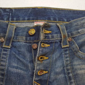 May include: Close-up of blue denim jeans with button fly. The jeans have a faded wash and visible stitching in yellow. The top button is gold with a logo. The label reads "TRUE RELIGION BRAND JEANS".