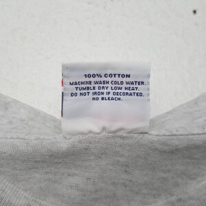May include: A close-up of a white tag with black text. The tag reads "100% COTTON, MACHINE WASH COLD WATER, TUMBLE DRY LOW HEAT, DO NOT IRON IF DECORATED, NO BLEACH." The tag is attached to a light gray fabric.