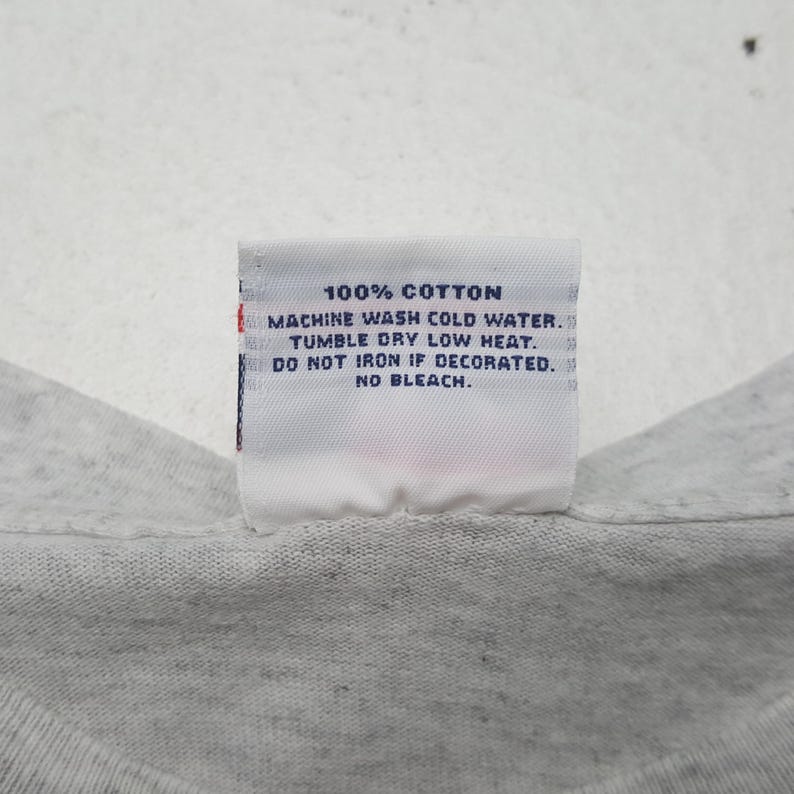 May include: A close-up of a white tag with black text. The tag reads "100% COTTON, MACHINE WASH COLD WATER, TUMBLE DRY LOW HEAT, DO NOT IRON IF DECORATED, NO BLEACH." The tag is attached to a light gray fabric.