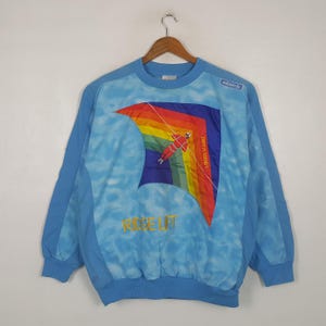 Puede incluir: Una sudadera de manga larga azul claro con un fondo de nubes y un gráfico grande de un ala delta arcoíris con un piloto. El texto "RIDGE LIFT" está bordado en amarillo y "THOSE FLYERS" aparece en el ala delta. Incluye el logotipo de Adidas.
