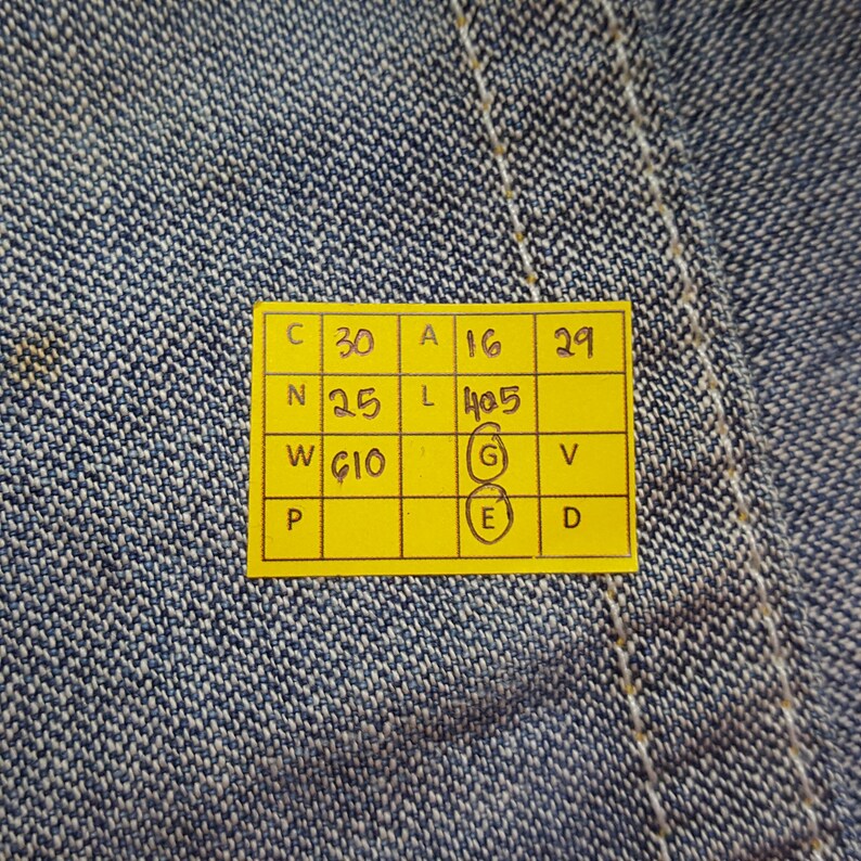 May include: Close-up of blue denim fabric with a yellow tag. The tag has several rows of numbers and letters, including "C 30", "A 16", and "W G10". The denim has visible stitching and a textured surface.