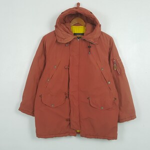 May include: A rust-colored winter coat with a hood, featuring a yellow lining visible at the collar. The coat has multiple pockets, buttons, and a zipper closure. A small zippered pocket is on the left sleeve.