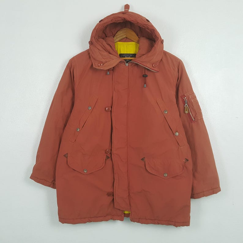 May include: A rust-colored winter coat with a hood, featuring a yellow lining visible at the collar. The coat has multiple pockets, buttons, and a zipper closure. A small zippered pocket is on the left sleeve.