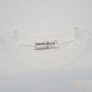 May include: White crew neck t-shirt with the text "SHARK ATTACK" on a tag near the collar. The tag also reads "100% COTTON MADE IN THAILAND". The shirt is made of a soft, comfortable material.