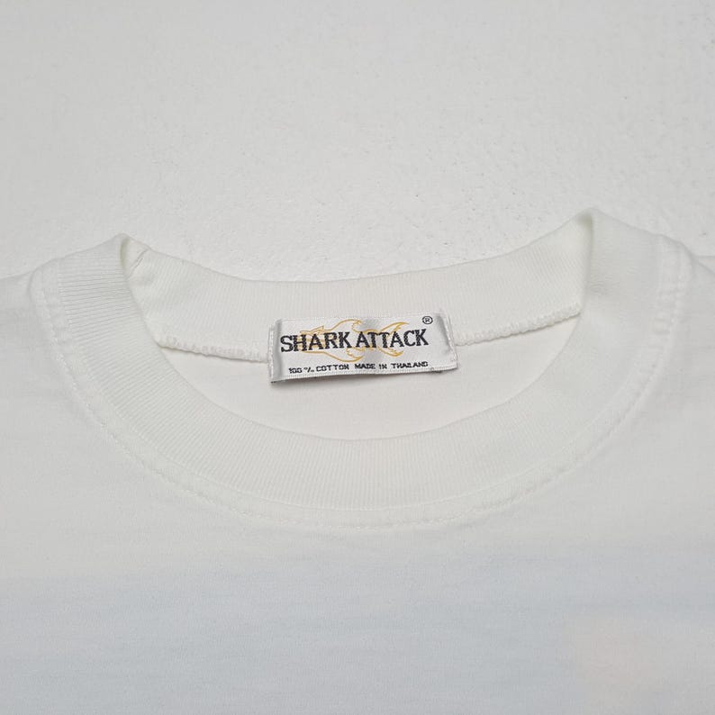 May include: White crew neck t-shirt with the text "SHARK ATTACK" on a tag near the collar. The tag also reads "100% COTTON MADE IN THAILAND". The shirt is made of a soft, comfortable material.