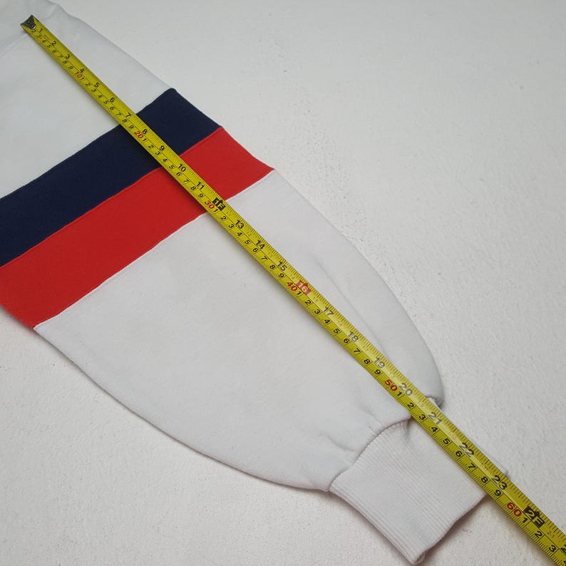 May include: White sweatshirt sleeve with a color block design featuring navy blue and red stripes. A yellow measuring tape is laid across the sleeve, showing measurements in inches.