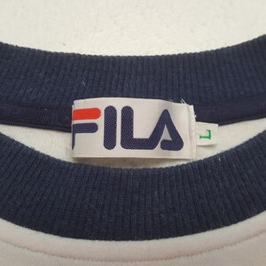 May include: Close-up of a white crew neck shirt with a navy blue collar and the brand name "FILA" in navy blue letters with a red bar above the "F" and "I". The shirt has a size "L" tag.