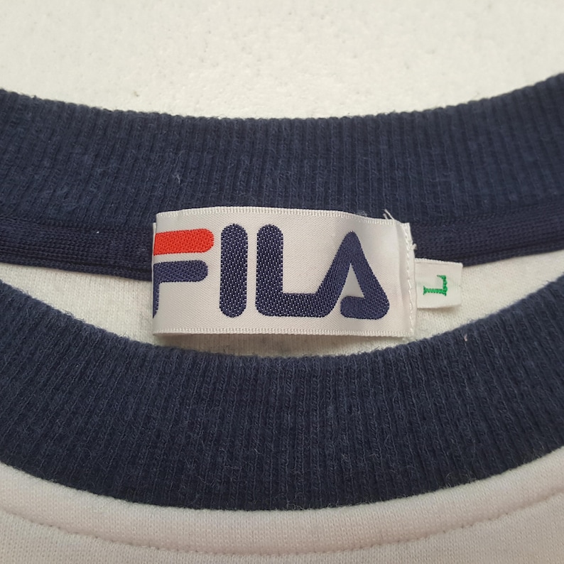 May include: Close-up of a white crew neck shirt with a navy blue collar and the brand name "FILA" in navy blue letters with a red bar above the "F" and "I". The shirt has a size "L" tag.