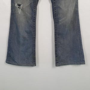 May include: Close-up of blue denim jeans with a distressed knee. The jeans have a faded wash and a slight flare at the bottom. The stitching is visible along the seams.
