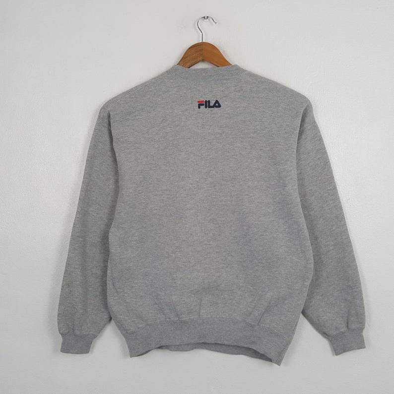 May include: A gray sweatshirt with long sleeves, hanging on a wooden hanger. The Fila logo is embroidered in red and navy blue on the upper back. The sweatshirt is made of a soft, comfortable material.